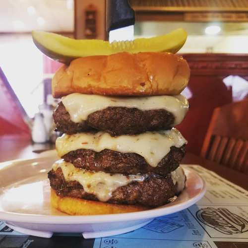 A towering burger with three patties, melted cheese, and a pickle on top, served on a plate in a diner setting.