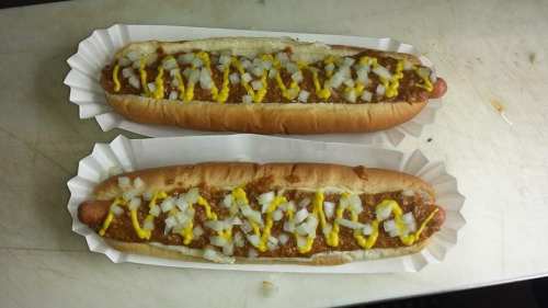 Two chili dogs topped with mustard and diced onions, served in paper trays on a countertop.