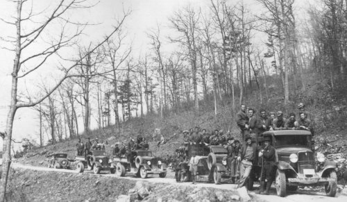 A historic black-and-white photo of a line of vintage cars with people gathered around them on a forested road.