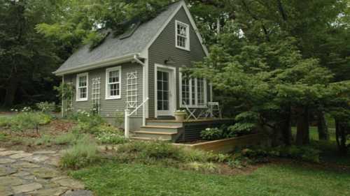 A small gray house with a porch, surrounded by greenery and trees, set in a peaceful outdoor environment.