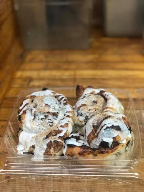 Two cinnamon rolls drizzled with icing, placed in a clear plastic container on a wooden surface.