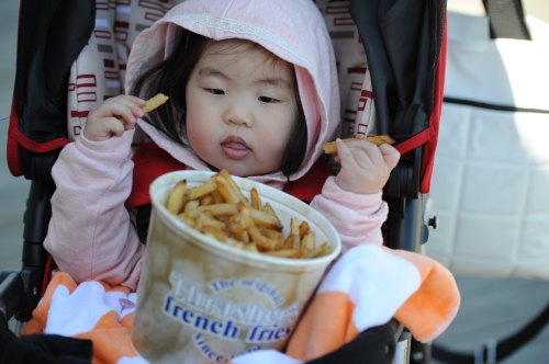 A young child in a stroller enjoys a large bowl of French fries, wearing a pink hoodie and looking curiously at the food.