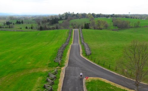 A cyclist rides on a road intersecting with a grassy field, surrounded by trees and wooden fences under a cloudy sky.