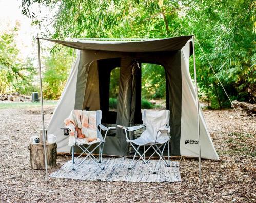 A cozy camping setup featuring a tent, two chairs, a blanket, and a small table in a wooded area.