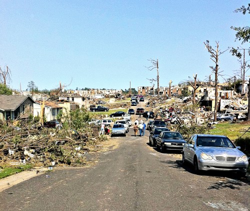 A street lined with damaged cars and debris, showcasing destruction from a recent tornado, with people walking amidst the wreckage.