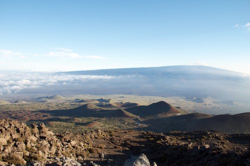 A panoramic view of a volcanic landscape with rolling hills, rocky terrain, and clouds beneath a clear blue sky.