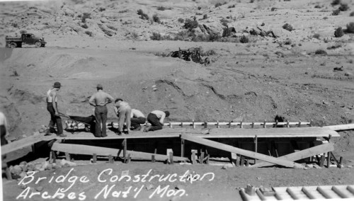 Black and white photo of workers constructing a bridge in a desert landscape, with vehicles and mountains in the background.