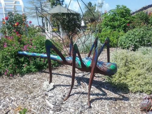 A colorful metal sculpture of a large grasshopper in a garden, surrounded by flowers and greenery.