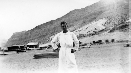 A man in a white outfit stands on a sandy area with mountains and structures in the background.