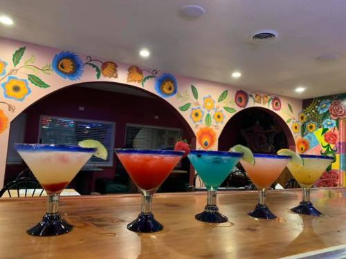 A colorful display of five cocktails in tall glasses, set against a vibrant floral mural in a lively bar.