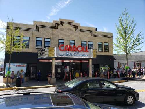 Historic theater building with "Carroll Arts Center" sign, people lined up outside, and trees lining the street.