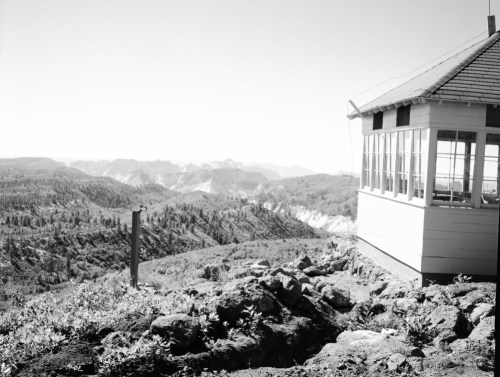 A lookout tower on a rocky hillside, overlooking a vast mountainous landscape in black and white.