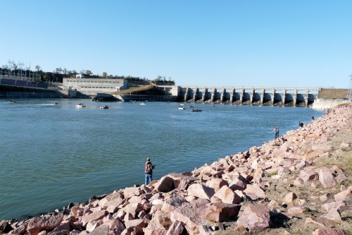 A river with a dam in the background, rocky shore, and several boats on the water. People are fishing along the bank.