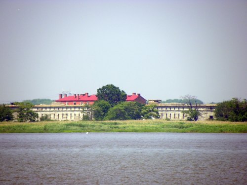 A historic building with a red roof, surrounded by greenery, viewed across a calm body of water.