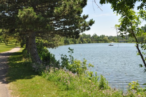 A serene lakeside scene with a path, lush greenery, and a small boat on the water under a clear blue sky.
