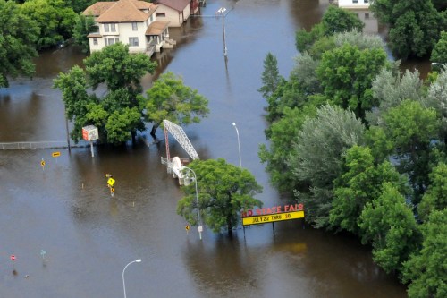 Aerial view of a flooded area with submerged trees, buildings, and a sign for a state fair partially underwater.