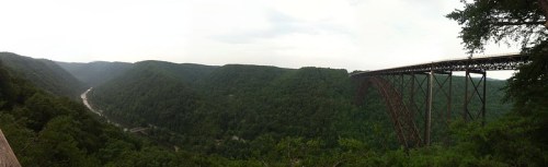 A panoramic view of a lush green valley with a tall bridge spanning across it, under a cloudy sky.