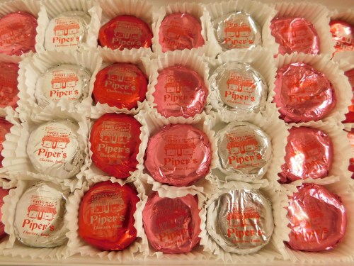 A box of assorted chocolate candies wrapped in red and silver foil, labeled "Piper's Chocolate Factory."