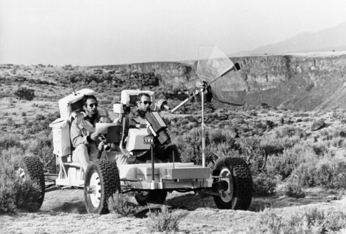 Two people drive a futuristic-looking vehicle across a rugged landscape, with mountains in the background.