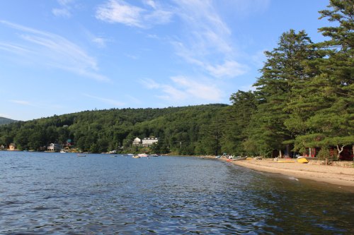 A serene lakeside view with a sandy beach, surrounded by lush green trees and distant buildings under a blue sky.