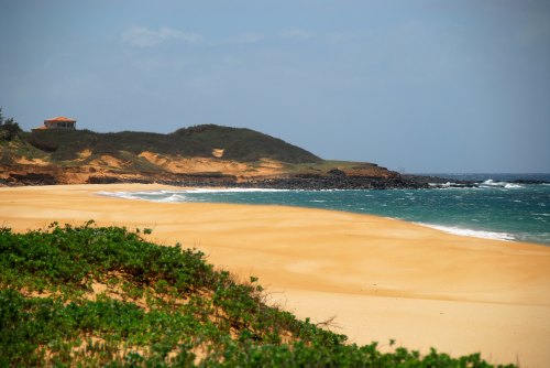 A serene beach with golden sand, gentle waves, and a distant green hill topped with a small house under a cloudy sky.