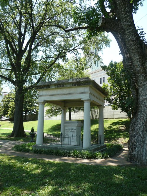 A stone monument with columns, surrounded by trees and grass, located in a park-like setting.