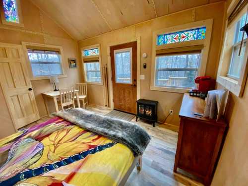 Cozy bedroom with colorful stained glass windows, a bed, small table, and a heater in a rustic setting.