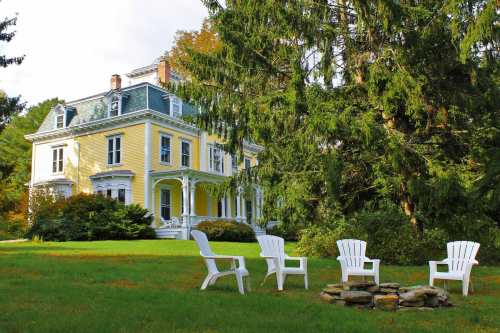 A large yellow Victorian house surrounded by trees, with white chairs arranged around a stone fire pit in the foreground.