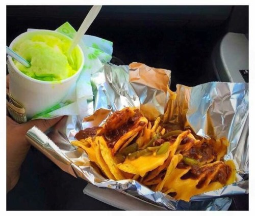 A hand holding a cup of green dessert next to a foil tray of nachos topped with sauce and jalapeños.