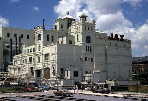 Historic brewery building with "Home of Jax Beer" sign, surrounded by cars and people, under a partly cloudy sky.