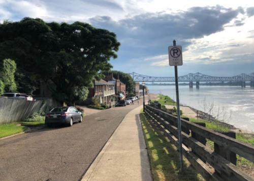 A riverside street with parked cars, trees, and a bridge in the background under a cloudy sky.