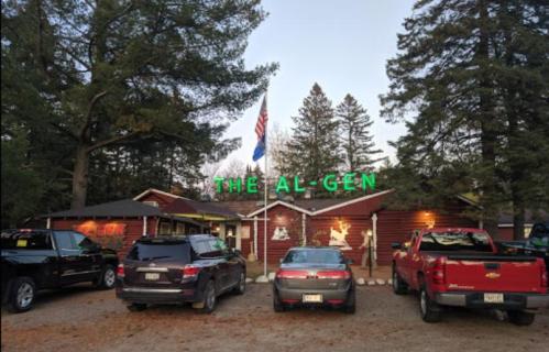 A rustic building with a neon sign reading "THE AL-GEN," surrounded by parked cars and tall trees.