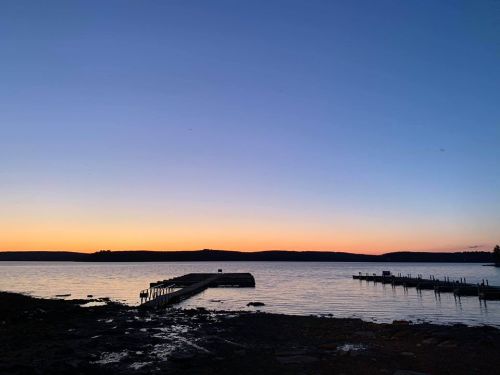 A serene lakeside view at sunset, with a dock extending into calm waters and a gradient sky of orange and blue.
