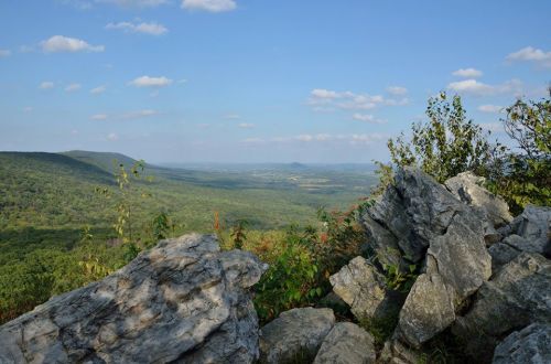 A scenic view of rolling green hills and valleys under a blue sky, framed by large rocks in the foreground.