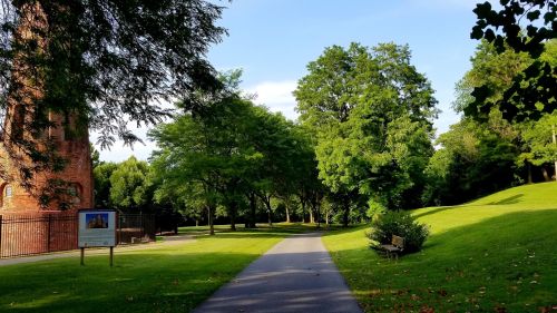 A peaceful park path lined with trees, leading to a grassy area and a historic brick building in the background.