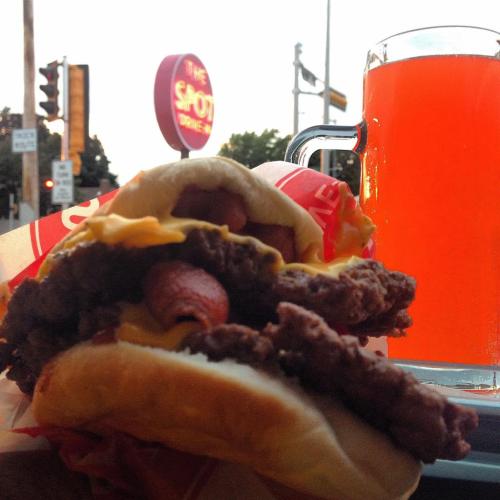 A close-up of a loaded burger with cheese and a hot dog, alongside a glass of bright orange drink.