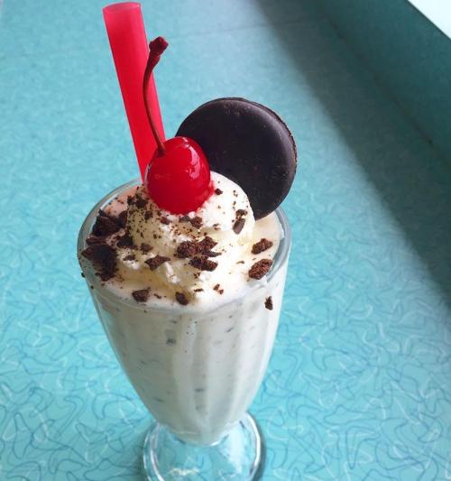 A tall glass of milkshake topped with whipped cream, a cherry, chocolate cookie, and crushed cookies, on a retro table.
