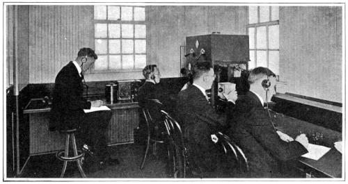 A historical image of four men in suits using early telecommunication equipment in an office setting.