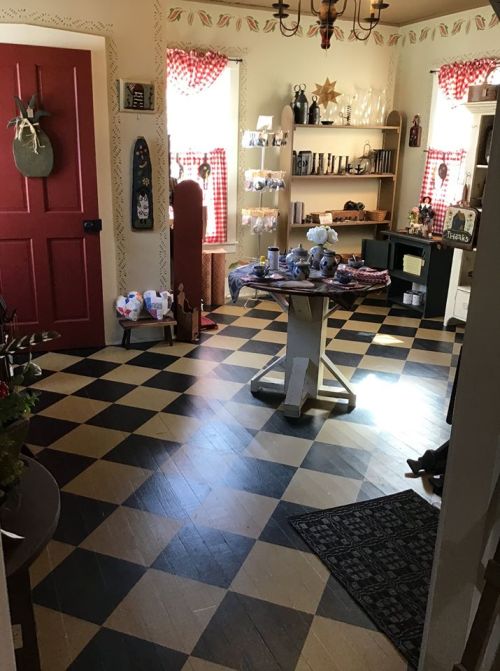 A cozy room with a checkered floor, red door, shelves of decor, and bright windows with checkered curtains.