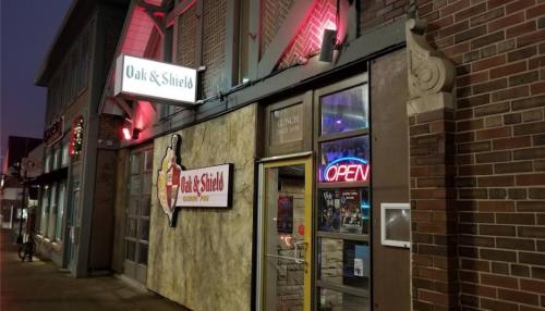 Exterior of Oak & Shield pub, featuring a lit "OPEN" sign and a rustic facade, set in an evening atmosphere.