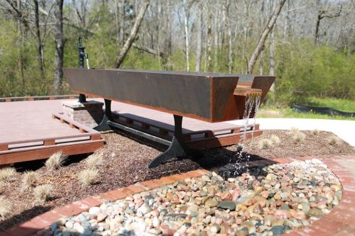 A modern metal water feature with a long basin, flowing water, surrounded by pebbles and greenery in a natural setting.