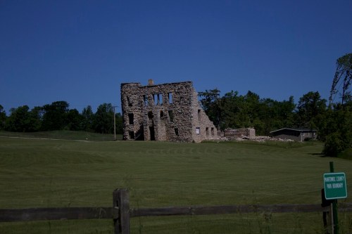 Ruins of a stone building on a grassy field, with trees and a clear blue sky in the background.