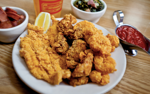 A plate of fried fish and chicken, served with lemon, dipping sauce, and sides of greens and sausage.