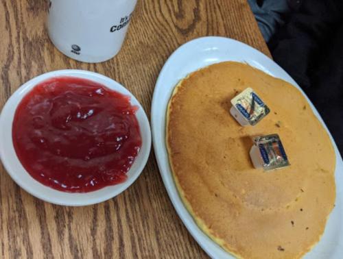 A large pancake with two butter pats on top, served with a small bowl of red fruit jam on a wooden table.