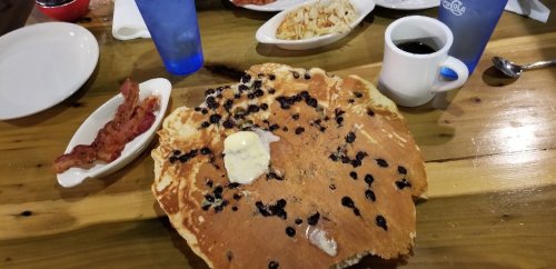 A large blueberry pancake with butter, served with bacon, coffee, and other breakfast items on a wooden table.