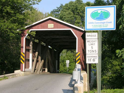 A red covered bridge over Little Conestoga Creek with signs indicating weight limit and clearance height.
