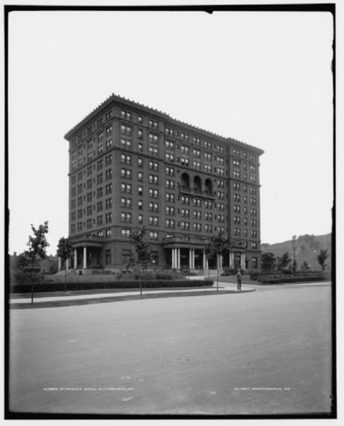 Historic hotel building with multiple stories, large windows, and a portico, surrounded by trees and a paved street.