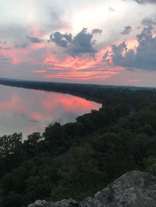 A serene sunset over a river, with vibrant pink and orange hues reflecting on the water and lush greenery along the shore.