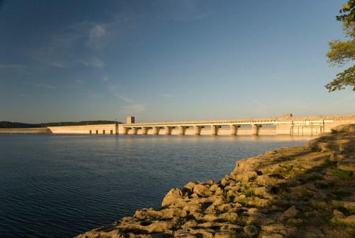 A serene view of a dam across a calm body of water, with rocky shoreline and clear blue sky.