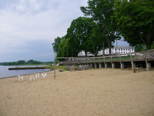 A sandy beach with white chairs, a wooden boardwalk, and trees along the shore under a cloudy sky.